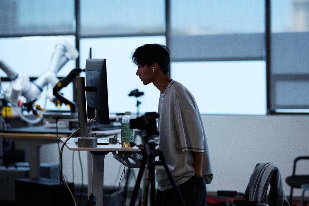 วิศวกรยืนตรวจสอบหน้าจอมอนิเตอร์ข้างแขนกลของ Generalist AI ในห้องทดลอง